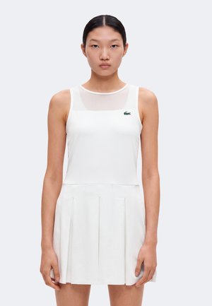 Femme portant une robe blanche sans manches avec jupe plissée et petit logo crocodile vert sur la poitrine, debout devant un fond clair uni.