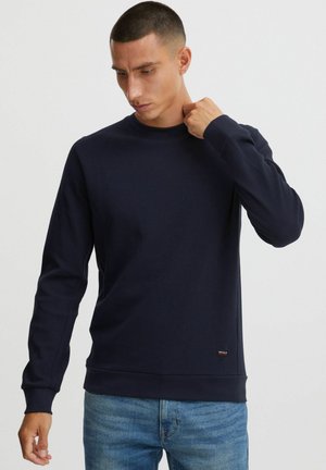Navy long-sleeve sweatshirt with a ribbed texture, round neckline, and contrasting cuffs. Small logo tag near the hem. Paired with light blue jeans.