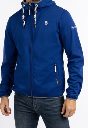 Homme portant une veste à capuche bleue zippée avec des cordons blancs et des poches zippées, associée à un jean bleu foncé.