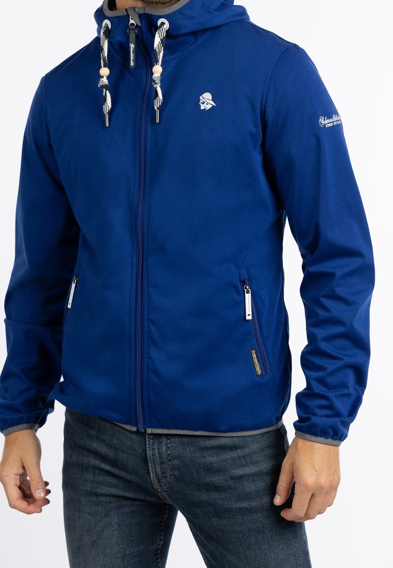 Homme portant une veste à capuche bleue zippée avec des cordons blancs et des poches zippées, associée à un jean bleu foncé.