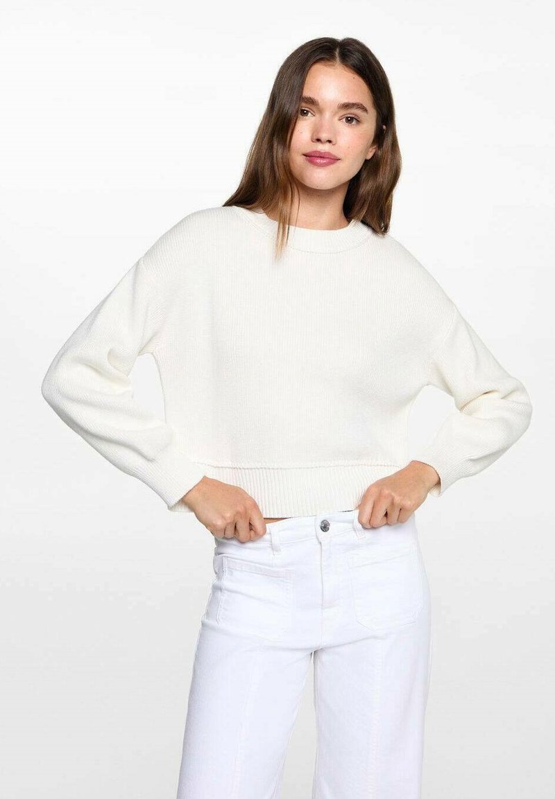 Weißer gerippter Pullover mit rundem Halsausschnitt und langen Ärmeln, ausgestattet mit einem cropped Schnitt und strukturiertem Strickdesign. Kombiniert mit weißen Jeans.