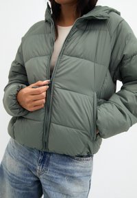 Polstret jakke i dæmpet grøn, med lynlås fortil, sidelommer og hætte. Stoffet fremstår glat og let blankt.