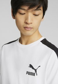 Camiseta blanca con acentos negros en los hombros, con un logo negro de Puma en el pecho. Material de algodón suave, diseño clásico de cuello redondo.