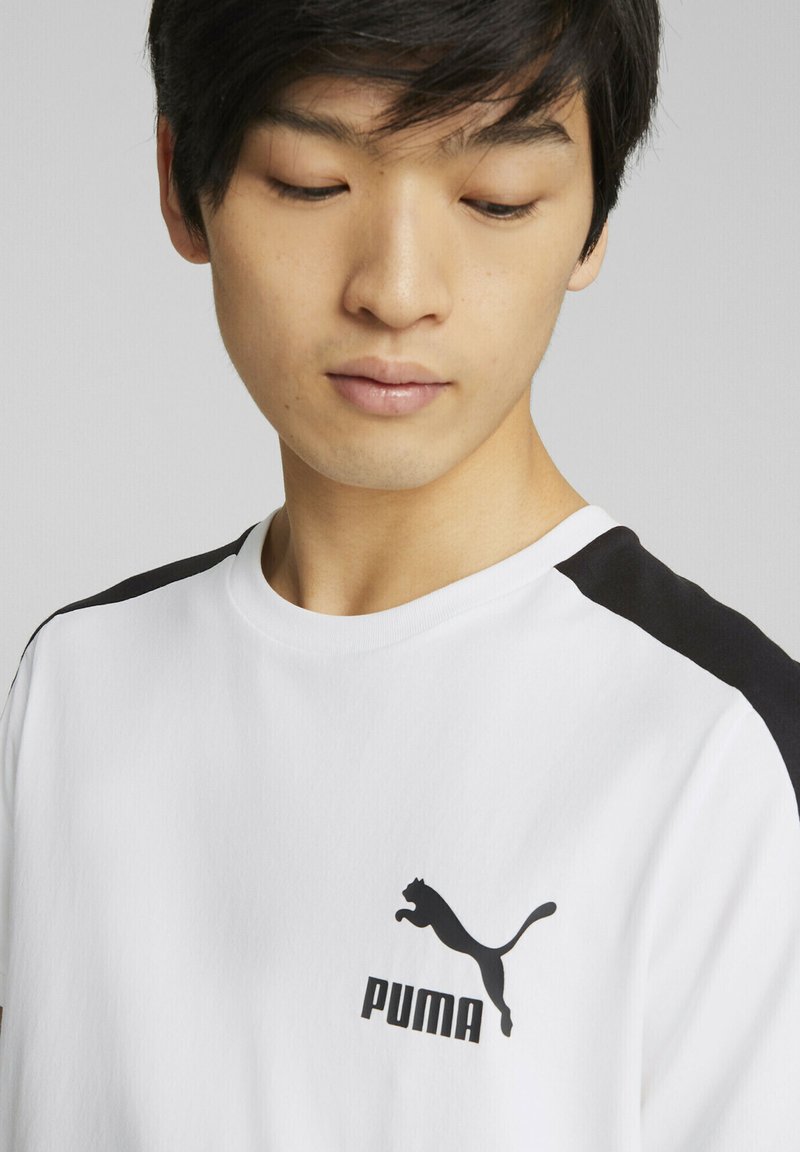 Camiseta blanca con acentos negros en los hombros, con un logo negro de Puma en el pecho. Material de algodón suave, diseño clásico de cuello redondo.