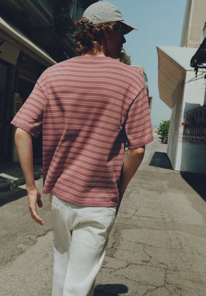 Personne portant une chemise rose à rayures, un pantalon blanc et une casquette grise marchant dans une rue urbaine ensoleillée avec des bâtiments de chaque côté.