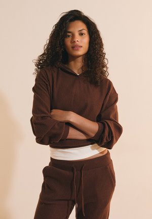 Woman with curly hair stands with arms crossed wearing a brown ribbed hoodie and matching pants against a plain beige background.