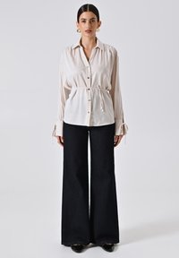 Camicia button-up beige chiaro con maniche lunghe e vita regolabile, abbinata a jeans a zampa scuri. Entrambi i capi presentano texture morbide.