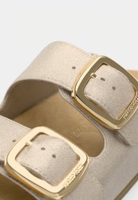 Κοντινό πλάνο από λυτές μπεζ λουριά σανδαλιών με δύο μεγάλες χρυσές ορθογώνιες αγκράφες και ορατές τρύπες ρύθμισης.