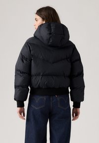 Casaco puffer preto com capuz, apresentando um design em losango, punhos canelados e um corte cropped. Usado sobre jeans de cintura alta azul escura.