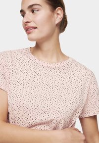 Mujer joven mirando hacia un lado, vistiendo una camiseta de manga corta rosa clara con pequeños lunares negros sobre un fondo liso.