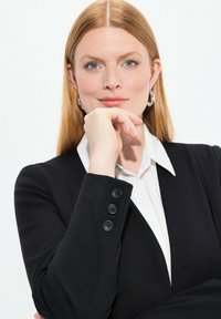 Blazer nero con dettagli ai polsi a tre bottoni, camicia bianca sotto e capelli rossi lisci di lunghezza media. Pelle chiara e occhi azzurri.