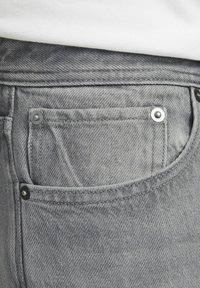 Primo piano della tasca di jeans in denim grigio con rivetti metallici, indossato con un top bianco.