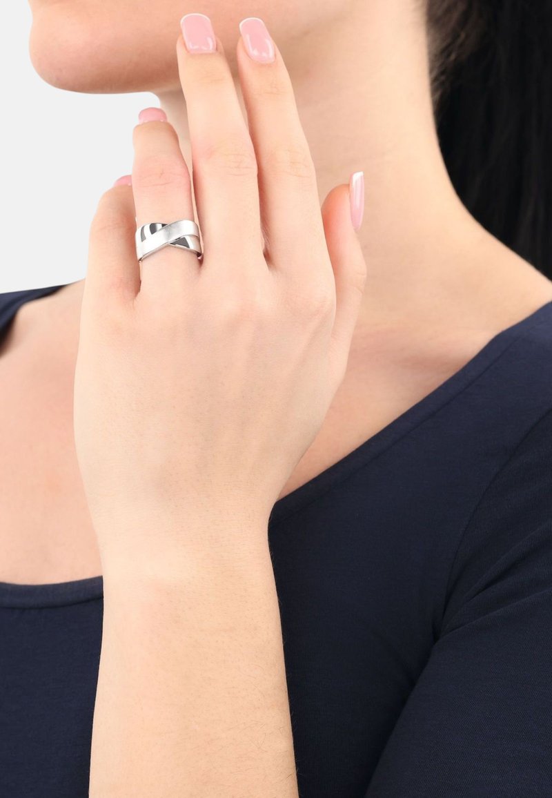 Anillo de metal plateado con una superficie lisa y pulida que presenta un diseño entrelazado, utilizado en una mano con uñas largas y de color claro.