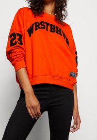 Sudadera naranja con letras negras "WRSTBHV" y el número "23" en las mangas. Puños y dobladillo acanalados, textura suave, etiqueta en el lado inferior.
