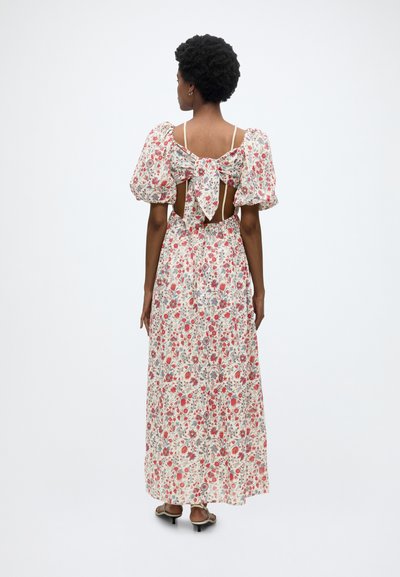 Femme aux cheveux courts et bouclés portant une longue robe fleurie avec manches bouffantes et un grand nœud au dos ouvert, debout sur fond blanc.