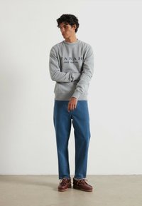 Grijze sweatshirt met "FARAH SINCE 1920 USA" in donkerblauwe tekst, gecombineerd met relaxed-fit blauwe spijkerbroek en bruine veterschoenen.