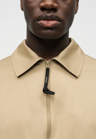 Κοντινό πλάνο ενός ατόμου με σκούρο χρώμα δέρματος που φοράει μια μπεζ ζακέτα με φερμουάρ και μαύρη λαβή φερμουάρ με την ένδειξη "ASPESI".