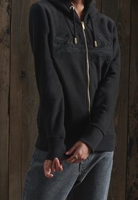 Felpa nera con zip realizzata in tessuto morbido, dotata di cappuccio, polsini a coste e un audace logo ricamato sul davanti, con dettagli della zip in oro.