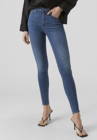 Smal blå denimjeans med hög midja, subtil blekning och förstärkt sömnad, tillsammans med svarta klacksandaler, mot en neutral bakgrund.