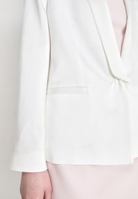 White blazer with a notched lapel, structured shoulders, and a single welt pocket. Smooth fabric with a subtle sheen.