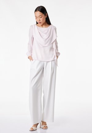 Vrouw staat met handen in de zakken, draagt een lichtroze gedrapeerde blouse, brede witte broek en witte sandalen met hoge hakken.