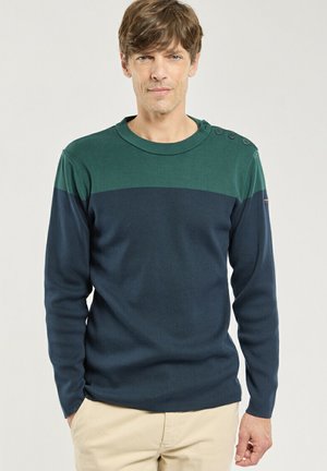Homme portant un pull à manches longues bleu foncé et vert avec des boutons sur l'épaule et un pantalon beige, debout devant un fond uni.