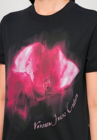 Black cotton t-shirt featuring a colorful pink floral graphic print and "Versace Jeans Couture" text in a shiny, contrasting font.