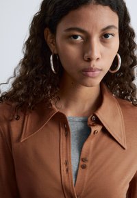 Brown button-up shirt with large collar, textured fabric, layered over a gray ribbed top. Silver hoop earrings are visible.