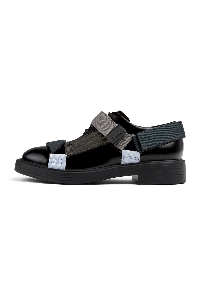 Zapatos de charol negro con correas elásticas en gris, azul y negro. Suela de goma gruesa, punta redonda y detalles de cierre ajustable.