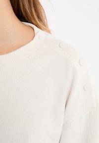 Jasnobeżowy prążkowany sweter z okrągłym dekoltem, z pięcioma białymi zatrzaskami wzdłuż ramienia. Miękka tekstura, casualowy design.