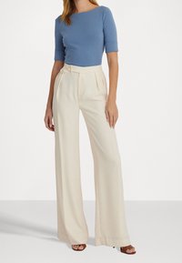 Donna con una maglietta azzurro a maniche corte, pantaloni alti a vita con gamba larga di colore crema e sandali con tacco aperti marroni su sfondo bianco.