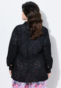 Blouse noire brodée avec un motif floral et des découpes, manches longues et poignets boutonnés, associée à un pantalon à motifs.