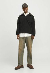 Schwarzer Fleece-Pullover mit Kragen, getragen über einem weißen Hemd. Kombiniert mit hellblauen Jeans und braunen Stiefeln. Eine schwarze Mütze rundet den Look ab.