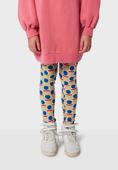 Lyserød oversized sweatshirt, mønstrede leggings med blå cirkler og røde accenter på et gult gitter, kombineret med hvide sneakers.