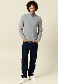 Suéter de punto gris con cuello alto y tres botones, combinado con jeans azul oscuro y zapatillas blancas. Presenta un patrón texturizado.