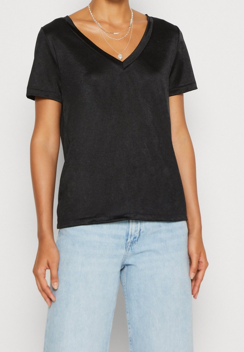 Personne portant un haut noir à manches courtes en V et un jean bleu clair, avec des colliers en argent superposés en accessoires.