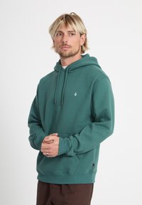 Hombre con cabello rubio, vestido con sudadera con capucha verde azulado y pantalones marrones, de pie con las manos entrelazadas, mirando ligeramente hacia un lado sobre un fondo blanco liso.