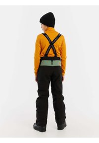 Pantaloni da sci con esterno nero, fascia in vita verde e cinturini regolabili. La persona indossa una maglia a maniche lunghe arancione e un berretto nero.