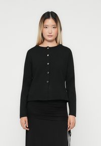 Woman with long straight hair wearing a black buttoned cardigan and black skirt, standing against a plain white background.