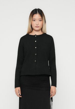 Woman with long straight hair wearing a black buttoned cardigan and black skirt, standing against a plain white background.
