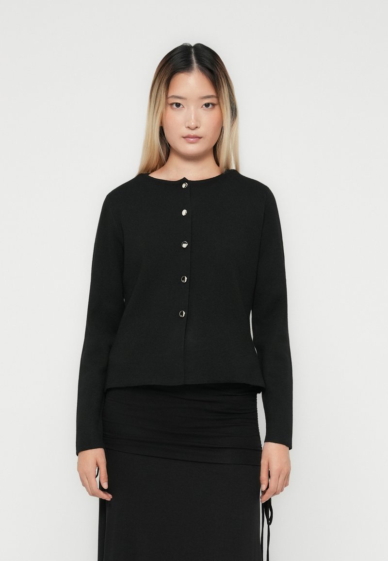Woman with long straight hair wearing a black buttoned cardigan and black skirt, standing against a plain white background.