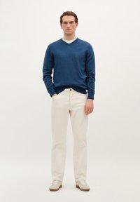 Maglione blu a scollo a V in morbido tessuto knit, abbinato a pantaloni bianchi a gamba dritta e sneakers beige. Il modello è in piedi di fronte.