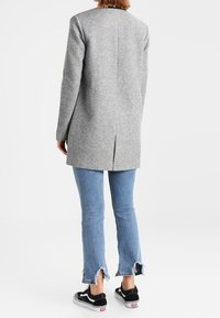 Cappotto lungo grigio in misto lana con taglio diritto e spacco posteriore, abbinato a jeans blu chiaro con orlo sfrangiato e sneakers nere.