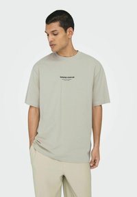 Lichtgrijze T-shirt met korte mouwen en een ronde halslijn, met zwarte tekst "CURATED STAPLES" op de voorkant. Zacht, casual materiaal met een relaxte pasvorm.