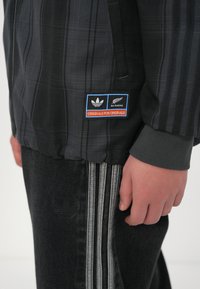 Casaco preto xadrez com fecho éclair e punhos canelados, apresentando um patch do logotipo da Adidas All Blacks. Combinado com calças de ganga escuras com detalhes em riscas brancas.