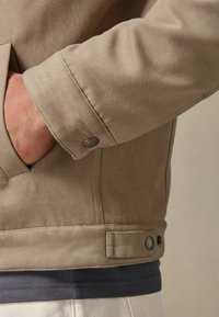 Mâneca unei jachete de suede bej cu detaliu de nasture, având un finisaj texturat și o cămașă stratificată albastră și gri dedesubt.