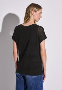 Svart stickad kortärmad topp med ett texturerat mönster, med rund halsringning och avslappnad passform. Bakvy som visar en semi-genomskinlig design.