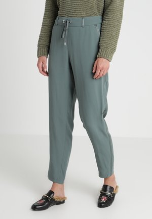 Person iført lysegrønne snorebukser, olivengrøn tekstureret sweater og sorte slip-on loafers med for af pels og blomsterbroderi.
