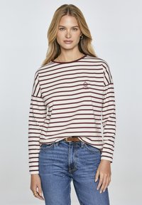 Long-sleeve striped shirt in cream and maroon with a round neckline and a small embroidered design on the chest, paired with blue jeans.