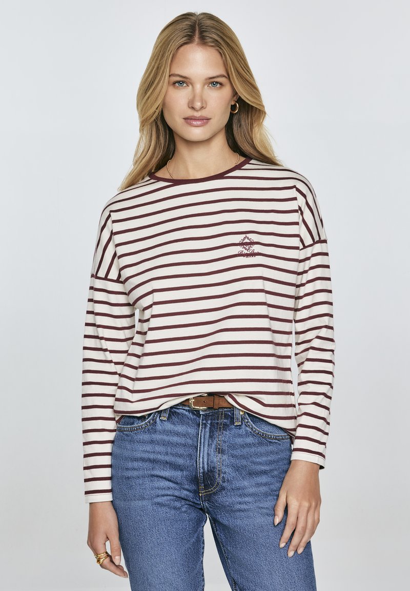 Long-sleeve striped shirt in cream and maroon with a round neckline and a small embroidered design on the chest, paired with blue jeans.